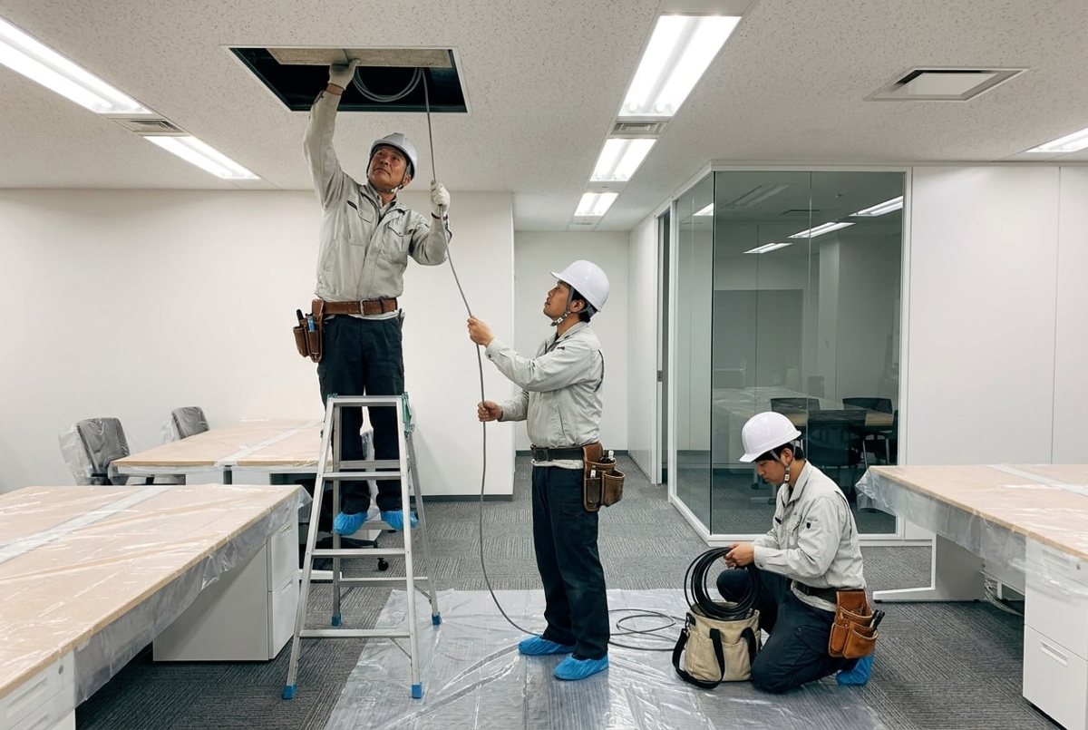 床にシートを敷き机にビニールをかけた事務所で、天井の点検口からケーブルを引く作業員